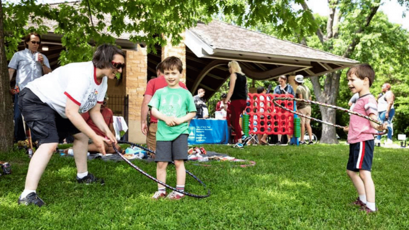 A parent holds a hula hoop around a small boy while another child hula hoops on his own in the grass with picnic games in the background in Saint Paul, MN.